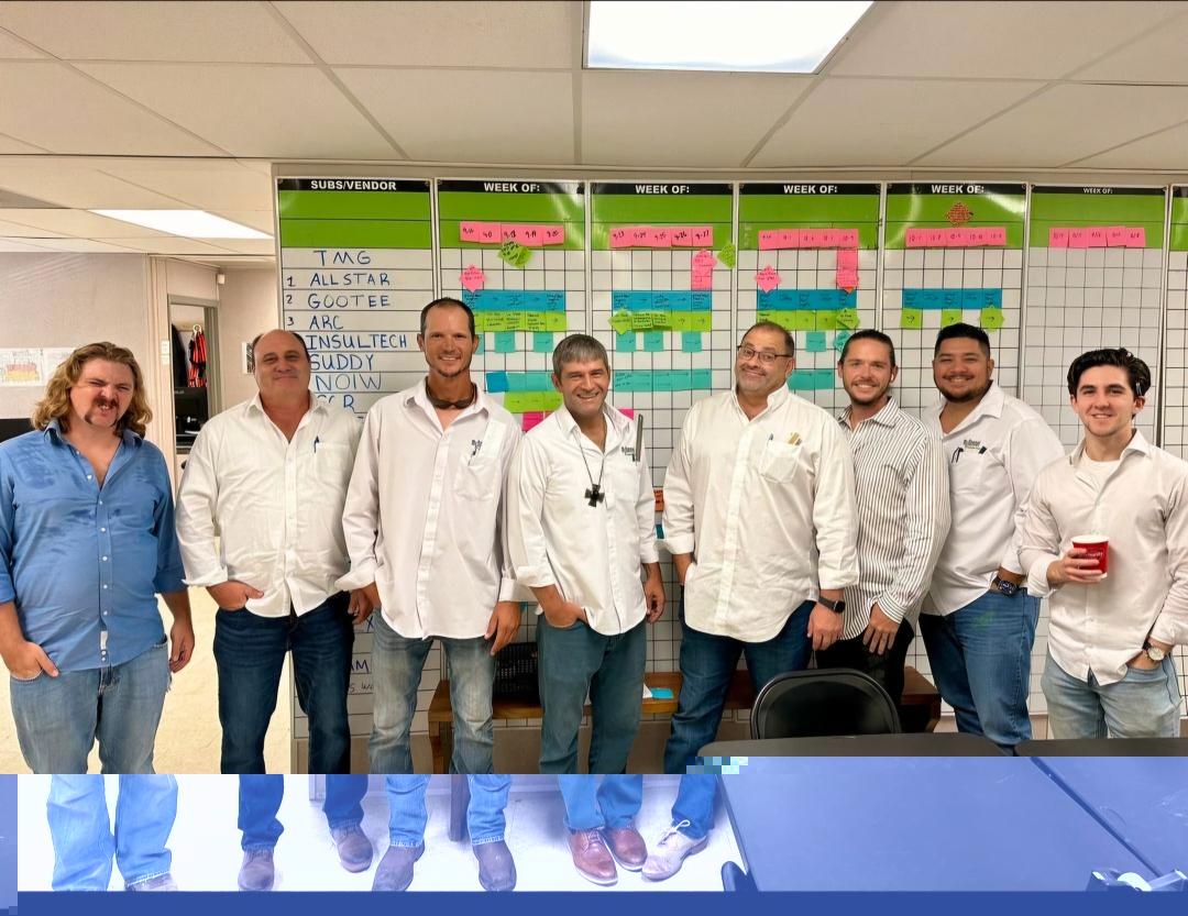 Group of McDonnel employees in white shirts smiling in front of a project planning board, showcasing teamwork and a supportive work environment.